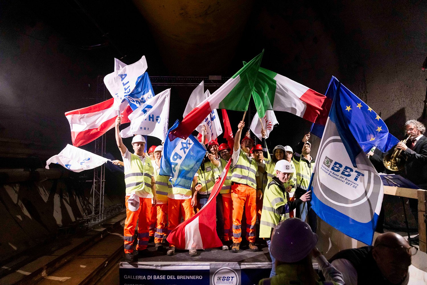 Ein Foto vom Abschluss des maschinellen Tunnelvortriebs in Italien. Im Tunnel stehen mehrere Menschen in Arbeitskleidung. Sie schwenken italienische und österreichische Flaggen. Zudem sind eine Europaflagge und mehre Fahnen mit Unternehmenslogos zu sehen.