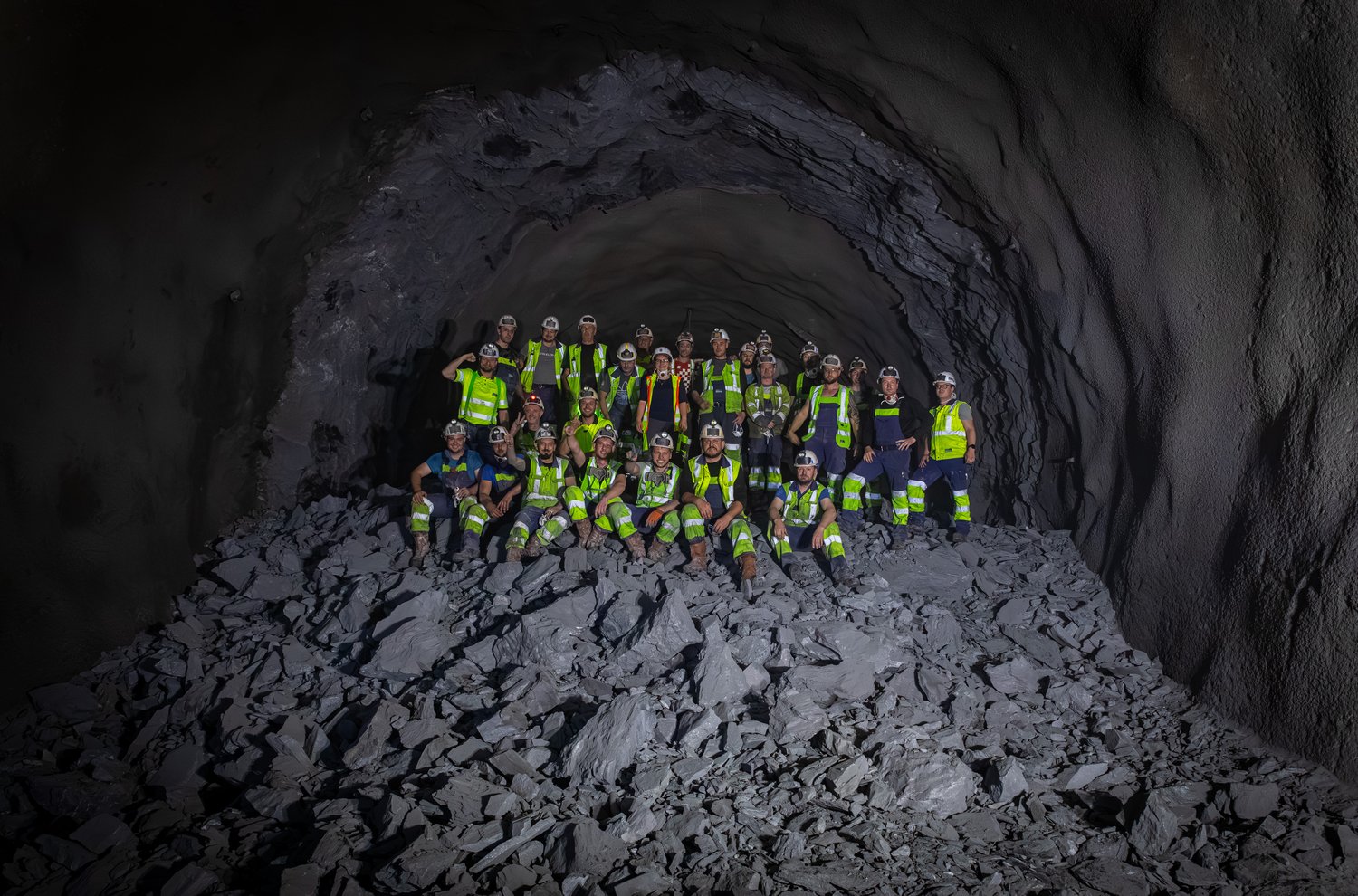Ein Foto von der Baustelle des Brenner-Basistunnels. 26 Arbeiterinnen und Arbeiter sitzen auf einem Haufen dunklen Gesteins. Sie tragen Helm und Arbeitskleidung und winken in die Kamera. Um sie herum sind die Wände des ausgebrochenen Tunnels zu sehen.