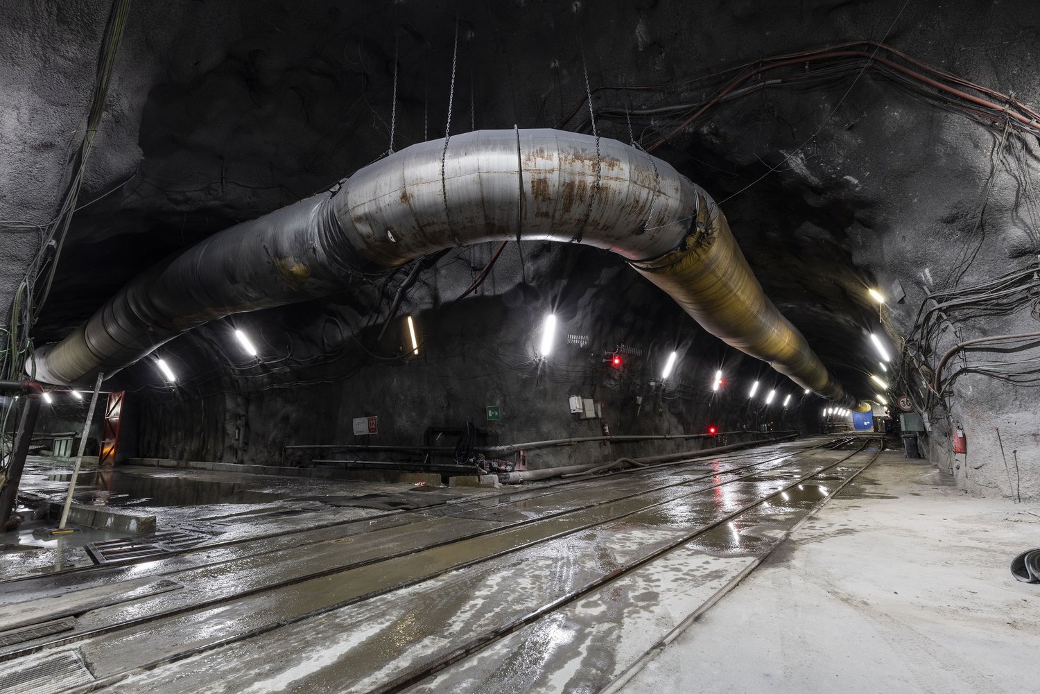Ein Foto von der Baustelle des Brenner-Basistunnels im Rohbau. Die Tunnelwände sind mit Spritzbeton gesichert. An der Decke ist der Schlauch einer Belüftungsanlage befestigt. An den Tunnelwänden sind Kabel, Schläuche und Leuchten angebracht.