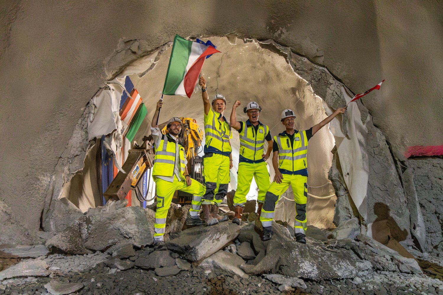 Vier Mineure stehen im durchgeschlagenen Erkundungsstollen. Sie stehen auf einem Haufen mit ausgebrochenen Gestein und schwenken die Flaggen Italiens, Österreichs und der Europäischen Union. Im Hintergrund ein Bagger mit Meisel.