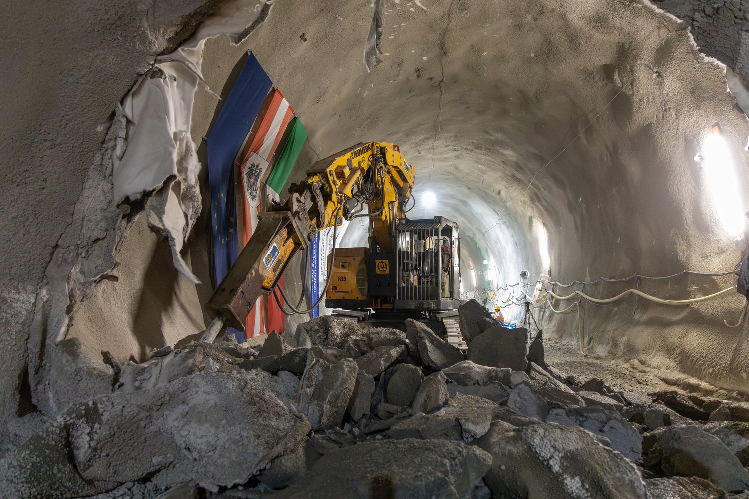 Ein Hydraulikbagger mit Meißel im durchgebrochenen Erkundungsstollen. Im Vordergrund liegt ausgebrochenes Gestein. An der Tunnelwand hängen die Flaggen der EU, der Republik Österreich und der Italienischen Republik.