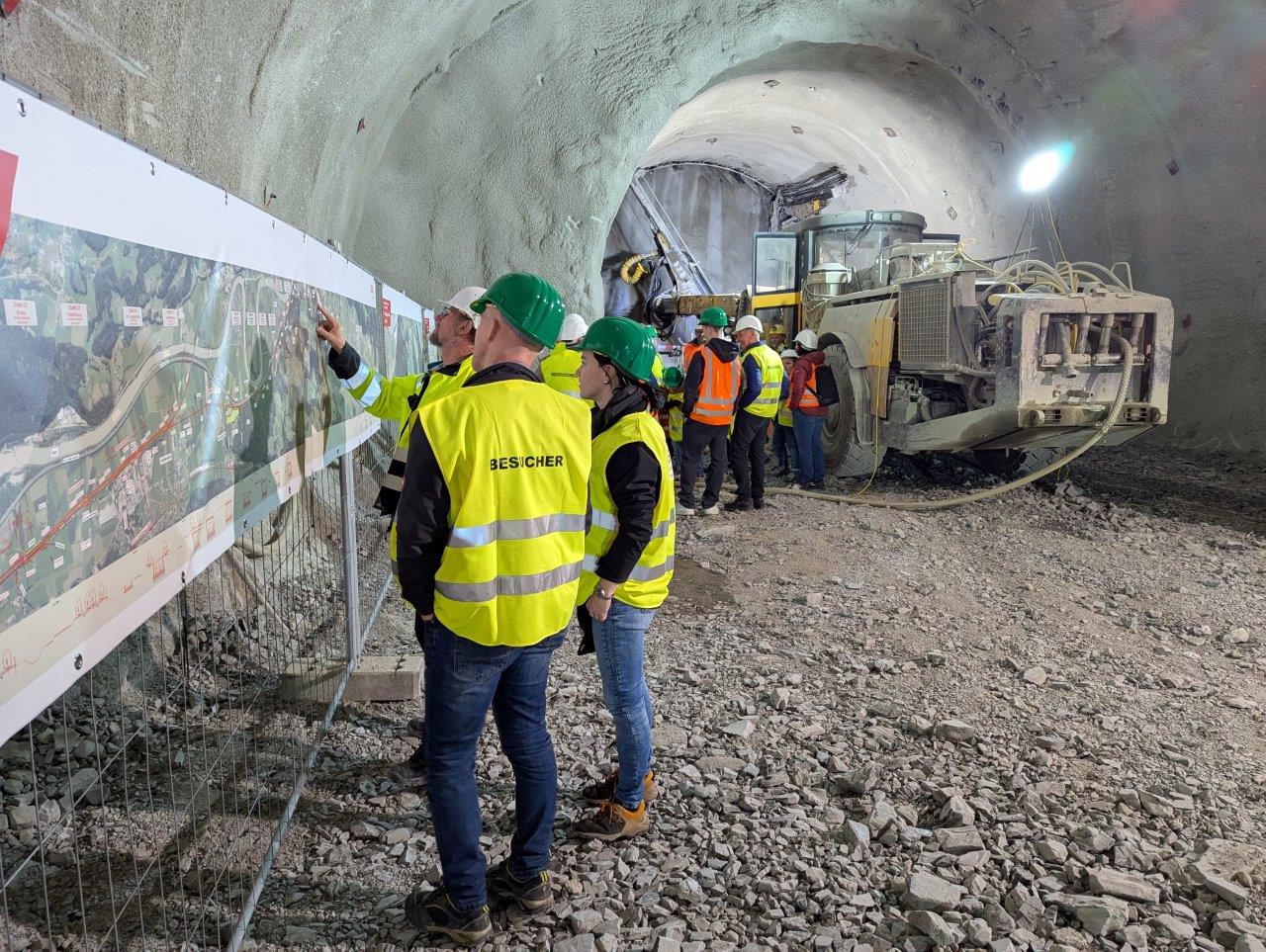 Eindrücke vom Besuch Anwohnender im Rohbaustollen Angath. Menschen mit Warnweste und Helm stehen im Tunnel und blicken auf Pläne der Neubaustrecke. Im Hintergrund steht direkt an der Ortsbrust eine große Baumaschine.