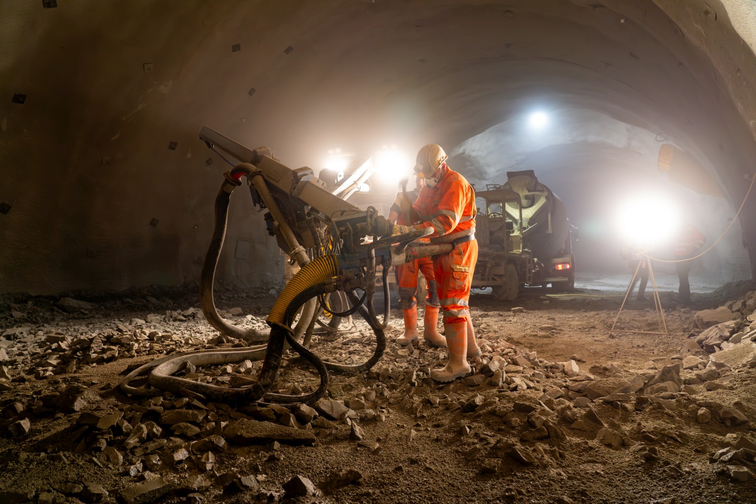 Ein Foto von der Baustelle des Rohbaustollens Angath. Im frisch ausgebrochenen Tunnel stehen spezielle Baumaschinen und ein Beton-Fahrmischer. Im Vordergrund arbeiten zwei Arbeiter in Schutzausrüstung an einem Spezialgerät.