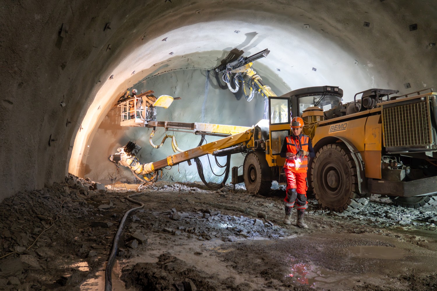 Ein spezielles Tunnelbohrgerät steht an der Ortsbrust. Auf einer Hebebühne arbeitet ein Bauarbeiter an der Tunnelwand. Zwei Arme der Maschine bohren in die Ortsbrust. Vor der Maschine steht eine Ingenieurin mit Schutzausrüstung und Helm.